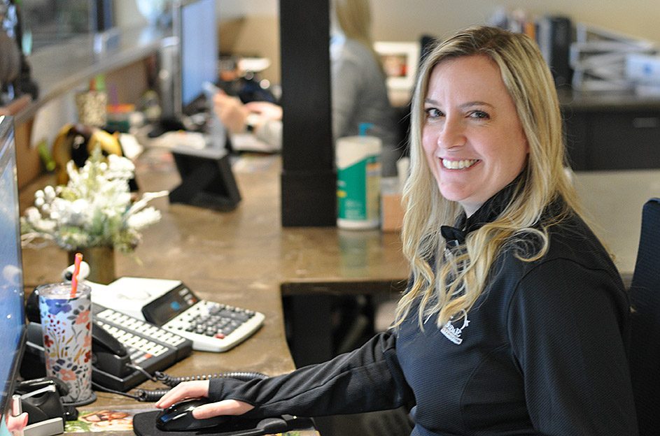 A woman with blonde hair is sitting at a desk, smiling at the camera. She is working on a computer and has office supplies, including a phone and a calculator, on her desk. In the background, other people are seated at their workstations.