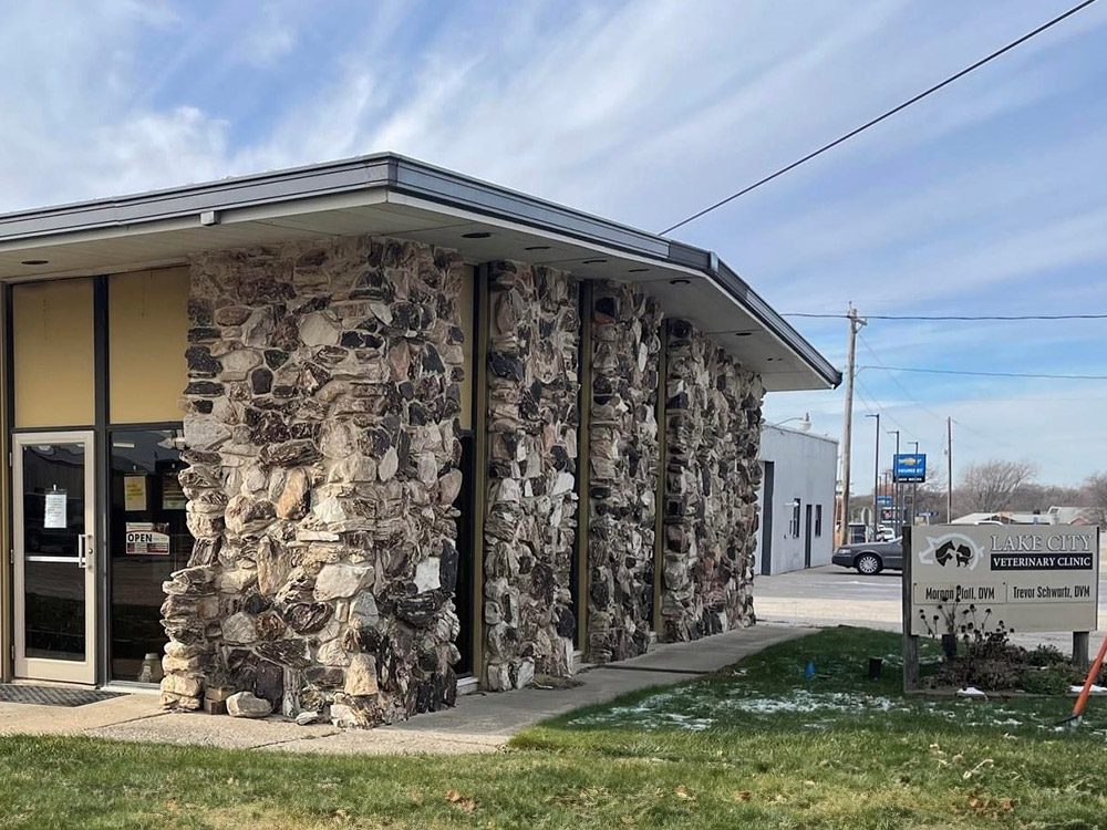 A one-story building with a stone exterior houses the "Mason City Veterinary Clinic." The entrance has glass doors. A sign displaying the clinic's name and contact details stands on the grassy area in front. The sky is partially cloudy, casting a gentle shadow over Lake City's IA-based clinic.