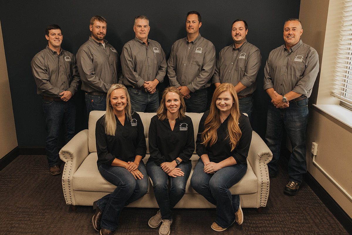 A group photo featuring six men standing in the back and three women seated on a couch in front, all dressed in matching gray shirts and jeans. The setting appears to be the professional office environment of Morris MN Clinic, with neutral-colored walls and a window on the right side.