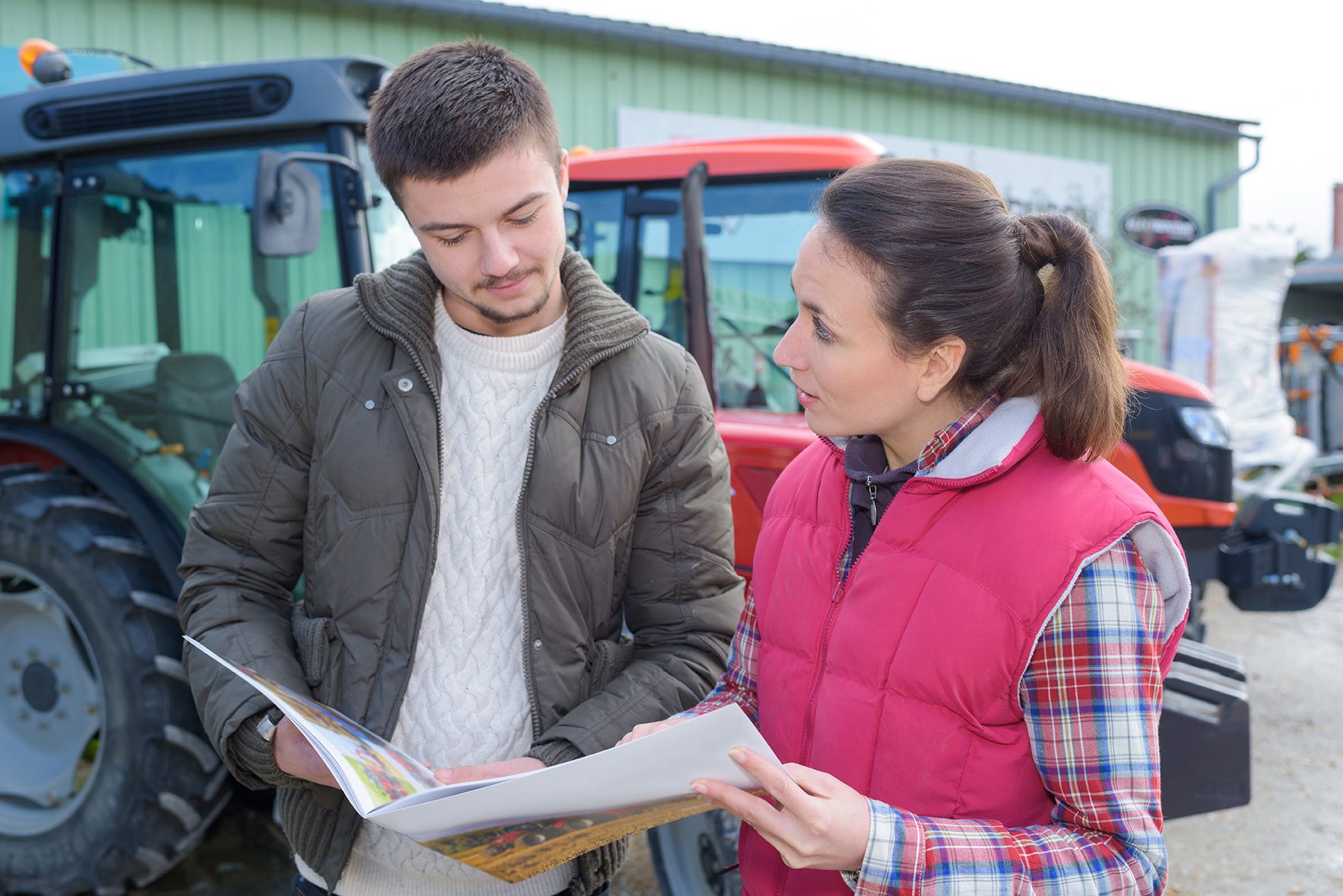 Two people are standing outdoors near farming equipment. One person, wearing a green jacket, is holding a booklet and looking at it. The other person, in a red vest and plaid shirt, appears to be explaining something, gesturing towards the booklet. .