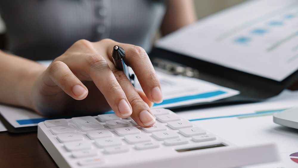 A person using a white calculator while holding a pen. Papers and documents are spread out on the table, and the person appears to be working on financial calculations or data analysis.