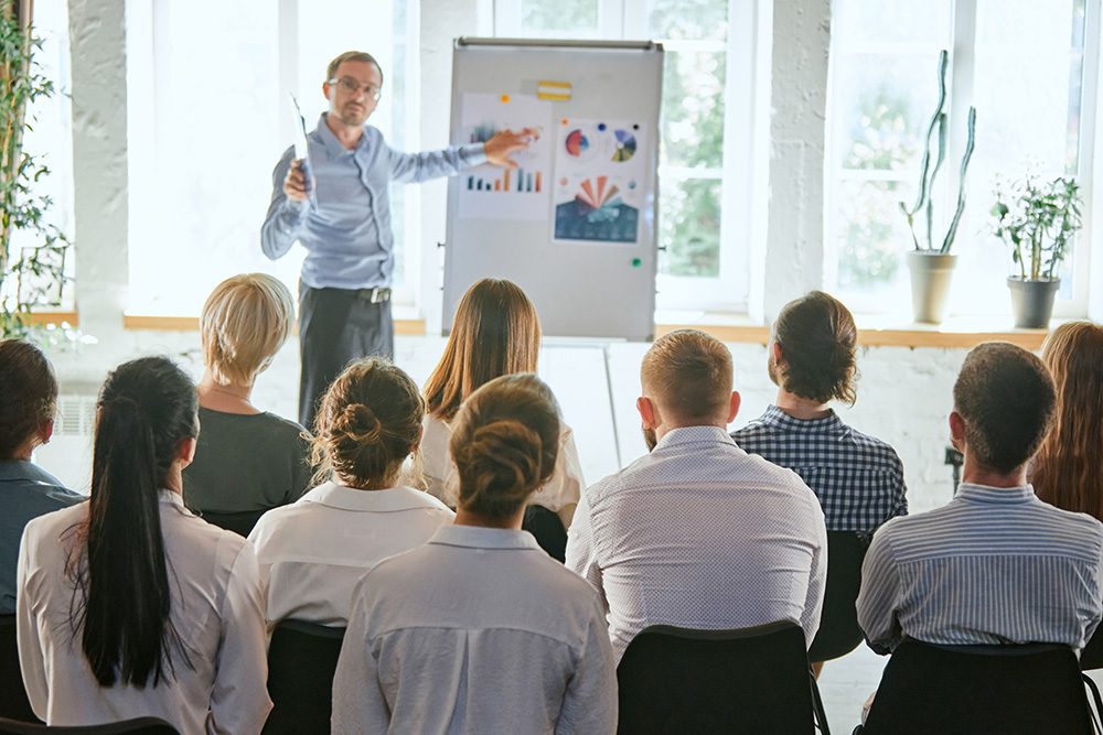A man is presenting to a group of seated people in a well-lit room. He points to a flip chart displaying various charts and graphs. The audience consists of both men and women, all facing the presenter. Potted plants are visible by the windows in the background.