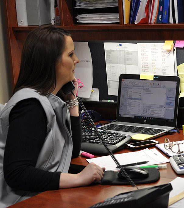 A woman sits at a desk, talking on a corded phone while looking at her computer screen, likely discussing product procurement. The desk is cluttered with papers, a keyboard, a mouse, and office supplies. Shelving with files and sticky notes is visible in the background.