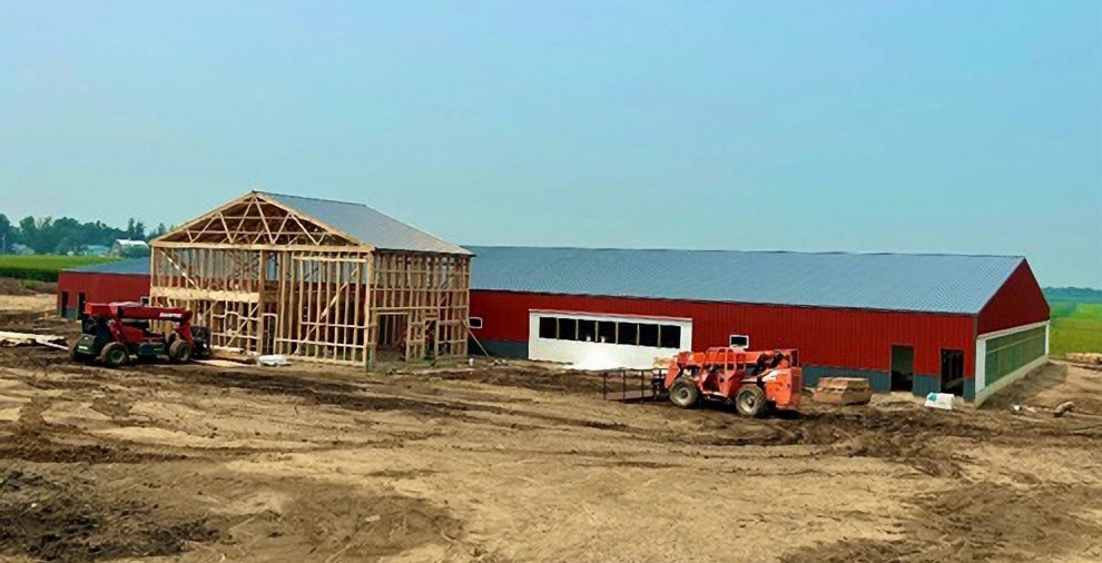 A large red barn is under construction in an open, dirt-covered area. As part of the project development, the building frame is partially completed, with two tractors and other construction equipment nearby. Fields of green crops are visible in the background under a clear blue sky.