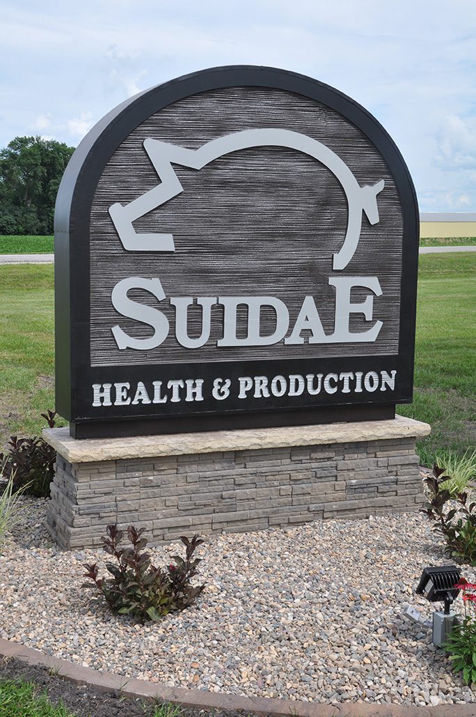A sign for “SUIDAE Health & Production Management” stands on a stone base with a gravel bed surrounded by plants. The logo features a stylized pig. The background consists of a grass field and trees under a partly cloudy sky.