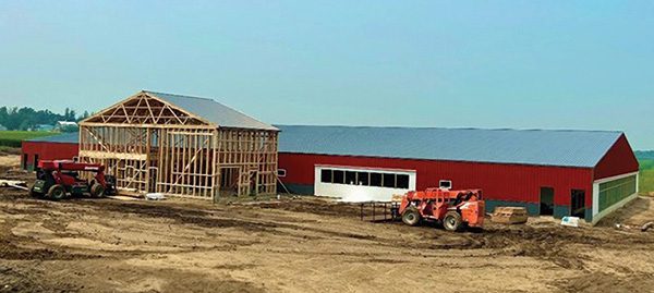 A large barn under construction in a rural area. The building has a partially completed wooden framework on the left, while the right side is fully built with red siding and a metal roof. Several construction vehicles providing services are parked around the site on dirt ground.