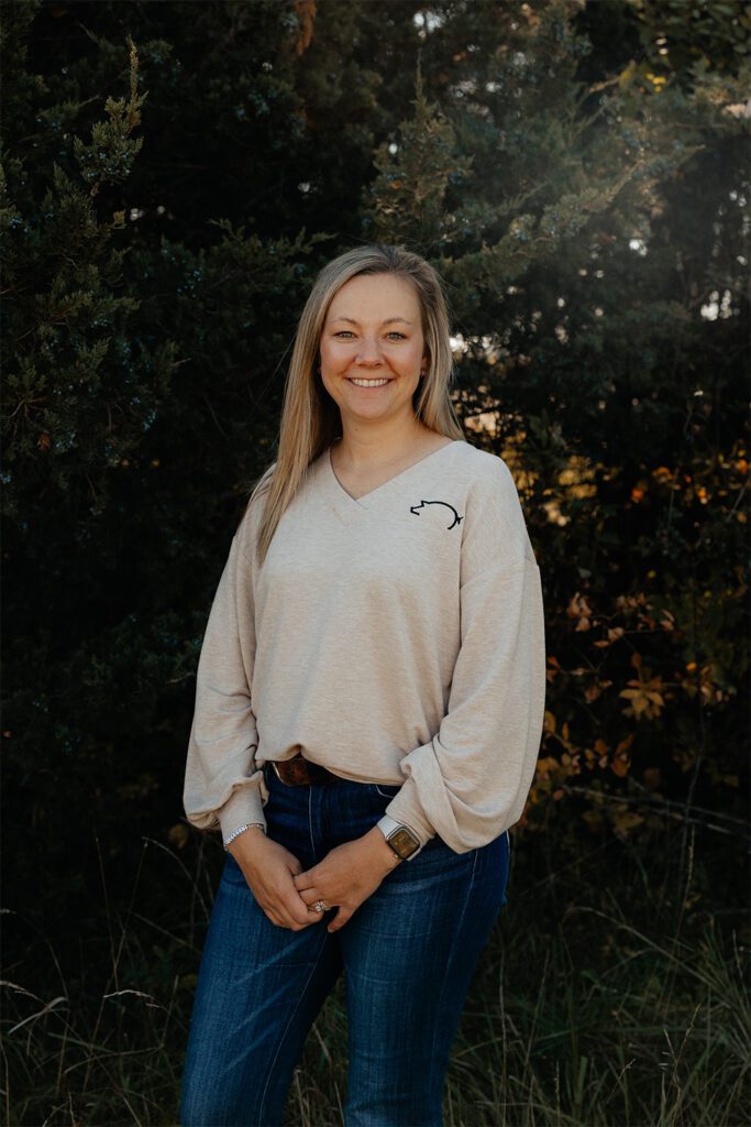 A woman with long blonde hair stands outdoors in front of leafy trees, smiling warmly. She is wearing a beige sweater with a small black outline of a horse on the shoulder and blue jeans. Her hands are clasped in front of her.