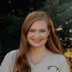 A woman with long red hair and fair skin smiles at the camera, wearing a light-colored V-neck shirt with a small pig outline logo. There are trees and sunlight in the background.