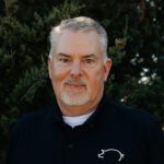 A middle-aged man with short gray hair and a goatee is wearing a black collared shirt with a white logo, standing outdoors in front of green foliage.