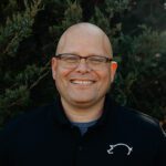A smiling bald man with glasses wearing a black collared shirt stands outdoors in front of green bushes.